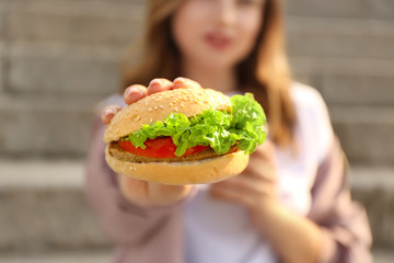 Beautiful young woman with tasty burger outdoors, closeup