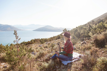 Woman traveler in autumn mountains above sea. Fall picnic with thermos of coffee or tea. Girl in plaid and hat is enjoying amazing view. Concept of traveling and relaxation at nature.