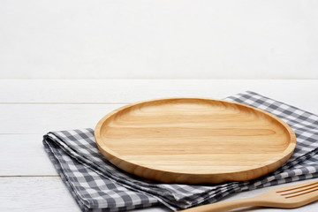 Empty round wooden plate with fork and grey gingham tablecloth on white wooden table.