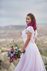 Fototapeta na wymiar Woman with a beautiful bouquet of flowers in her hands stands on the mountain in the rays of the dawn sunset. Beautiful white long dress on the girl body. Perfect bride with pink hair
