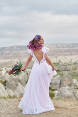 Fototapeta na wymiar Woman with a beautiful bouquet of flowers in her hands dance on the mountain in the rays of the dawn sunset. Beautiful white long dress on the girl body. Perfect bride with pink hair dance