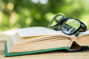 Open book and eyeglasses on a wooden table in a garden. Sunny summer day, reading in a vacation concept