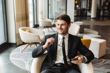 Photo of joyful young man sitting in hotel hall with smartphone and eyeglasses during business trip