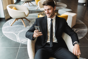 Photo of beautiful young man sitting in hotel hall with smartphone during business trip