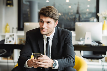Portrait of brunette young businessman sitting on armchair with smartphone in hotel hall