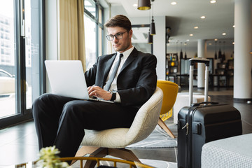 Portrait of caucasian young businessman sitting on armchair with laptop computer and suitcase in hotel hall