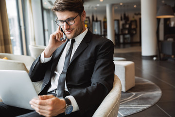 Portrait of attractive businessman using laptop computer and talking on smartphone in hotel hall