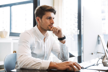 Image of serious businesslike man sitting at table and working on computer in office
