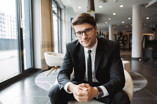 Photo of young businessman wearing eyeglasses sitiing on armchair in hotel lobby