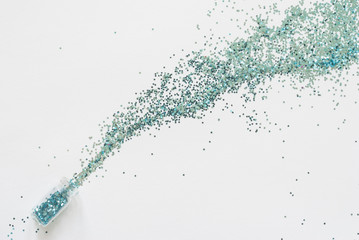 Turquoise blue glitter confetti spill out of a small glass jar diagonally on a white paper background.