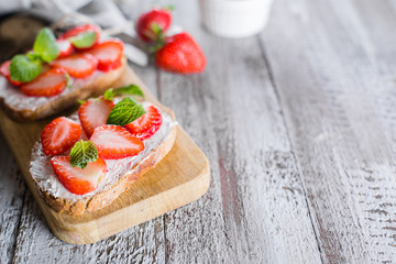 Two toasts or bruschetta with strawberry and mint on cream-cheese on wooden board on table background. Copy space