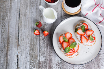 Two toasts or bruschetta with strawberry and mint on cream-cheese and cup of coffee on wooden table background. Top view, copy space