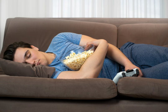 young handsome man fall asleep after playing fascinating video game still holding joystick in one hand and popcorn in other