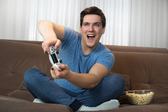 young handsome man playing video game sitting on comfy sofa holding joystick with both hands looking involved in mission