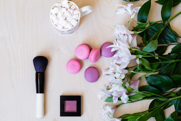 Obraz na płótnie Canvas Flat lay of macaroons, flowers, cup of cacao with marshmallow and makeup brush, pink blush. Wooden background. Copy space, mockup, top view, overhead