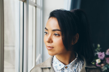 beautiful Asian woman with a hairstyle in Japanese style and a kimono at the window on the background of the home interior