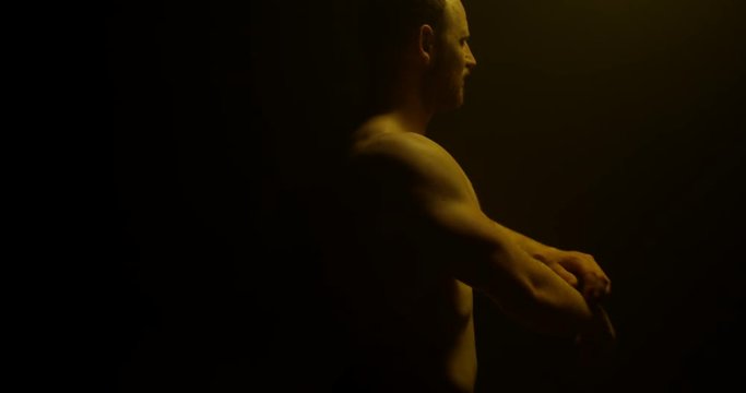 Male Body Builder Preparing For Session In A Dark Room, Training For Body Building Competition. Dim Lit Room, Yellow Light Over Him. Training Hard Preparing For Competition