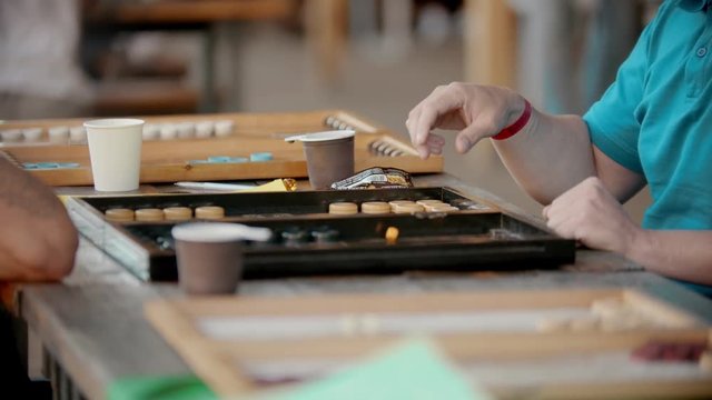 Two men playing backgammon at the table outdoors - one man throwing a dice - two cups are next to them