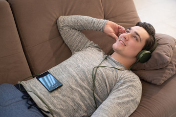 Obraz premium young handsome smiling man lying on the sofa listening to music in earphones from smartphone feeling relaxed