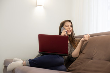 young beautiful smiling brunette girl sitting on the sofa talking on the phone and watching movie on laptop