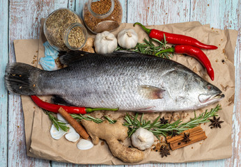 Fresh Raw Sea Bass on wooden table surrounded by fresh ingredients and spices.