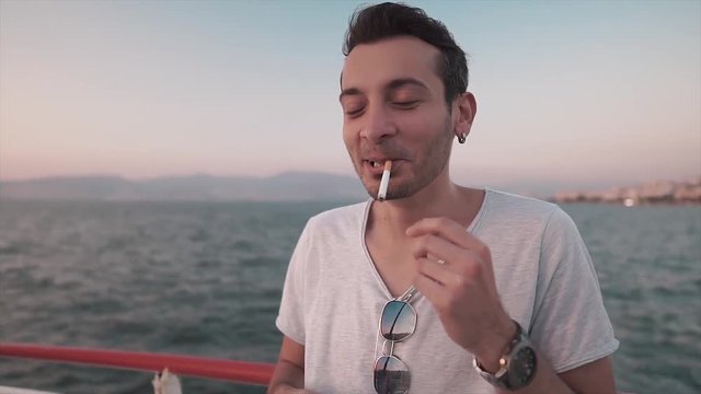 Handsome brunette young man is lighting cigarette near sea. Hipster guy smokes and laughs.