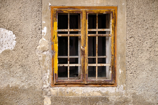 Minimalist capture of a single rural window on a grey textured wall with peeling concrete.  Simple window with yellow frame and mullions