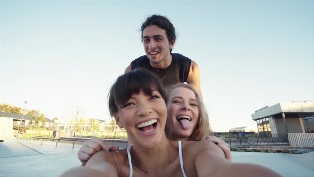 Man pushing female friends sitting on a skateboard and taking a selfie video. Group of friends creating a fun video at skateboard park.