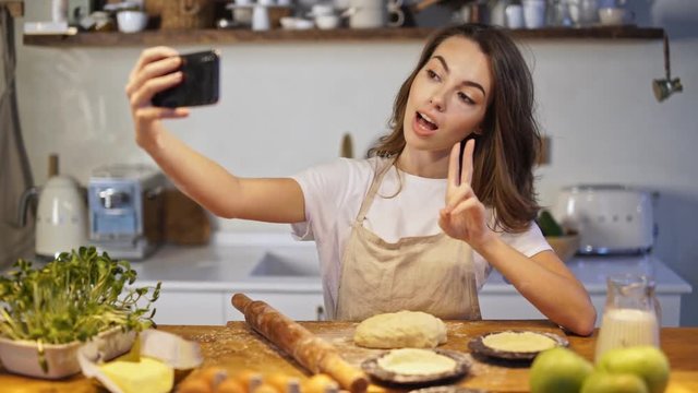 Attractive young woman in apron taking selfie photo on smartphone while cooking at the kitchen