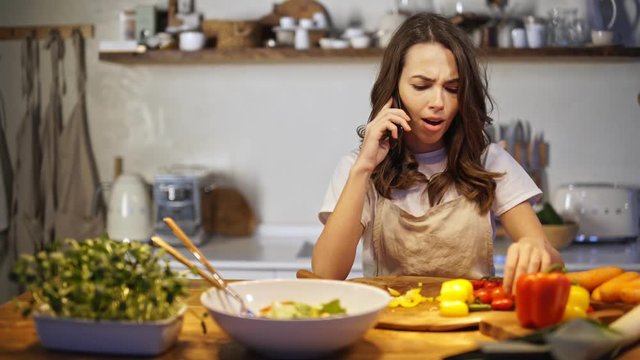 Cheerful young woman in apron talking on a smartphone while cooking in the kitchen