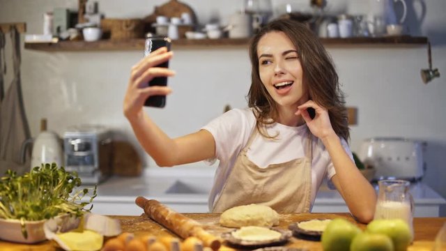 Smiling pretty lady in apron taking selfie photo on smartphone while cooking at the kitchen