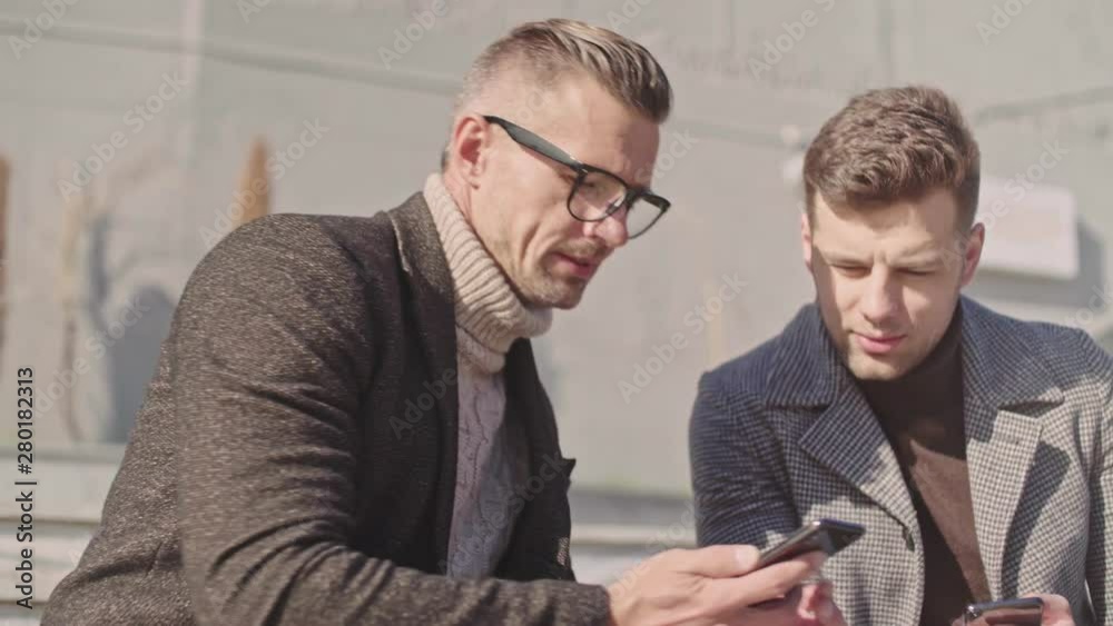 Poster two beautiful adult men communicate using smartphones while sitting outside