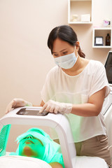 Asian young cosmetologist in protective mask turning on the special equipment with ultraviolet light while young woman getting beauty procedure