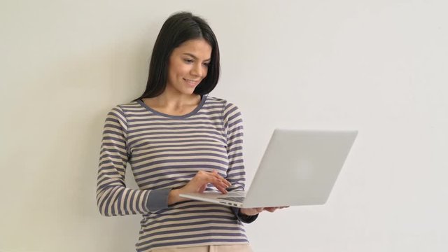 Attractive young woman texting on the laptop standing by the wall