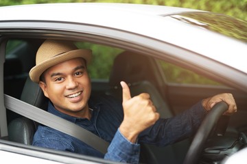 Portrait of happy smiling young asian man with fasten seat belts for safety travel on the road showing thumbs up while driving in his car.