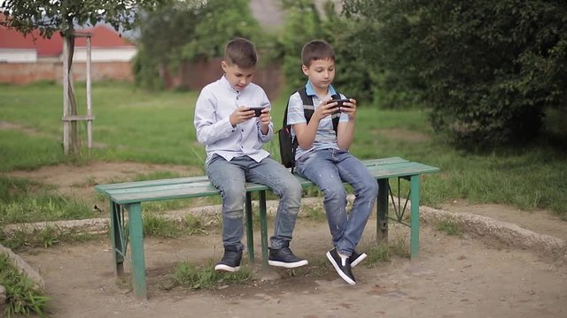 Two boy sitting on the bench and play online games. One boys with backpack. Young boys use their phones