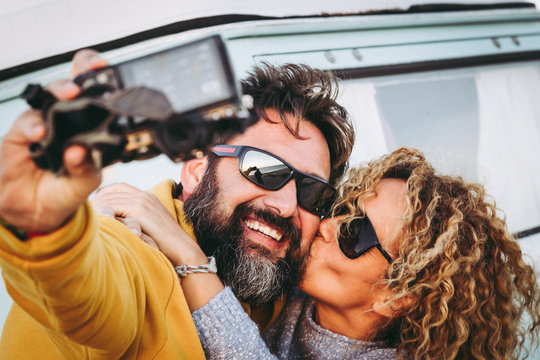Happy kissing couple taking selfie together  with vintage van in background. Husband kiss her wife making social video with camera to share online with friends. Trends lifestyle, travel love concept.