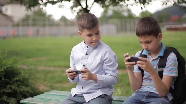 Two boy sitting on the bench and play online games. One boys with backpack. Young boys use their phones