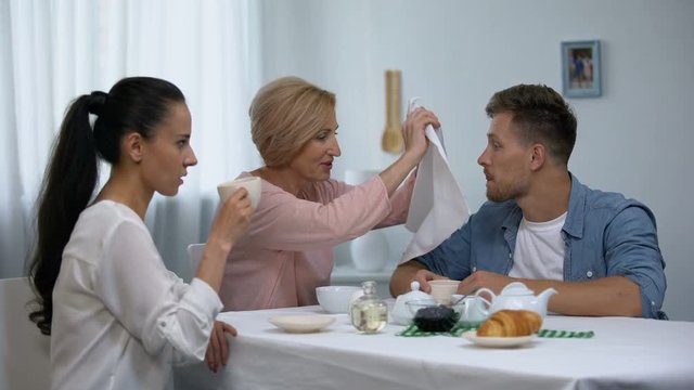 Shocked wife looking at mother-in-law putting napkin on husbands neck, tea party