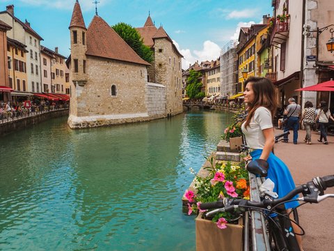 Brunette girl with long hair and blue skirt stands on the canal promenade in Annecy in France, everywhere flowers and bicycles