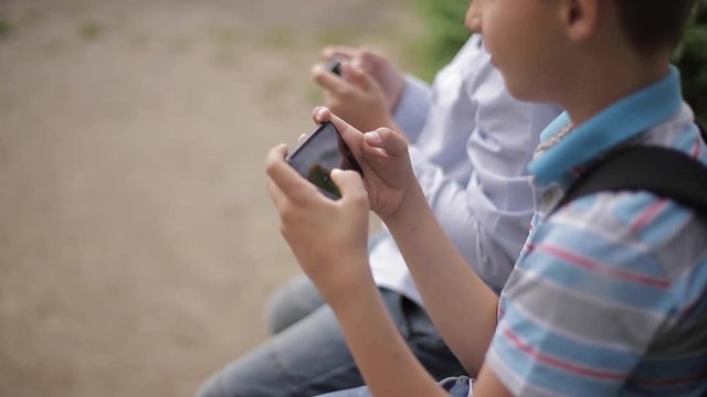 Two boy sitting on the bench and play online games. One boys with backpack. Young boys use their phones