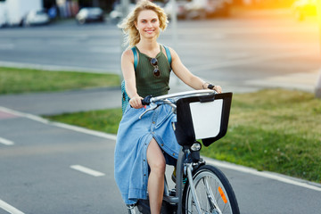 Photo of happy blonde in long denim skirt riding bike on road in city on summer day