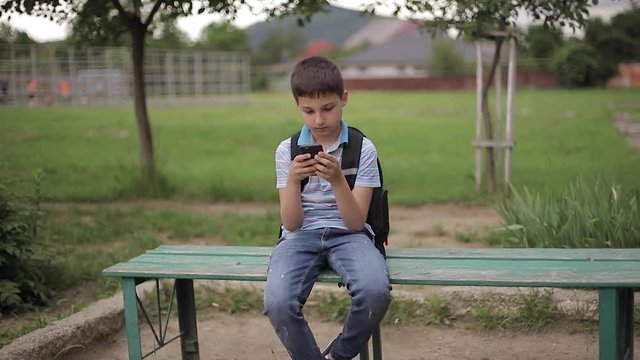 Young boy with backpack sitting on the bench and play online games during school break