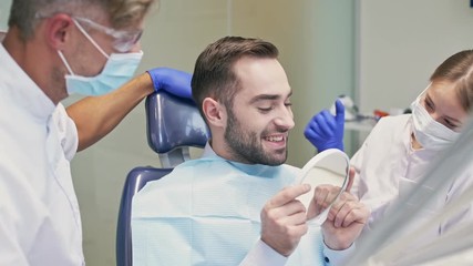 Beuatiful brunette man smiling and looking at the mirror after dental procedure in the dental clinic