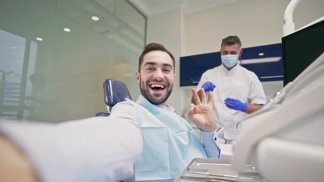 Smiling brunette man taking a selfie photo while dentist preparing and trying to start dental procedure at the dental clinic