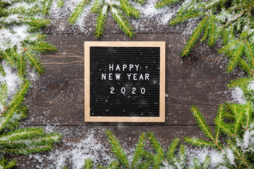 Christmas or new year frame for your project. Happy new year 2020 words on a letter board with Christmas fir tree branches and snow on a dark wooden board.