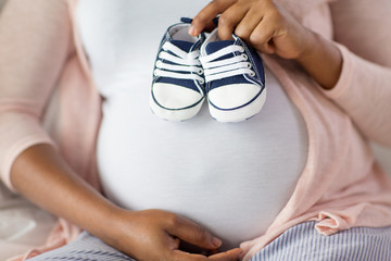 pregnancy, people and rest concept - close up of pregnant african american woman sitting on bed and holding little baby bootees