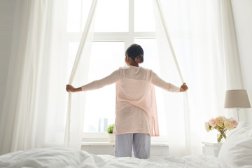 morning and people concept - happy african american woman opening window curtains at home bedroom