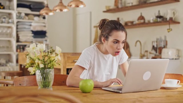 Cute young brunette woman in white t-shirt playing game on laptop computer and making winner gesture at the dining room