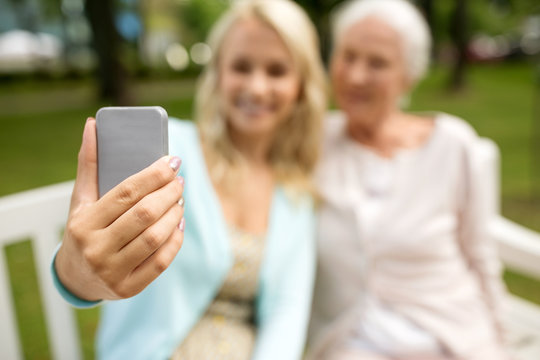 family, technology and people concept - close up of happy smiling adult daughter and senior mother with smartphone sitting on park bench and taking selfie